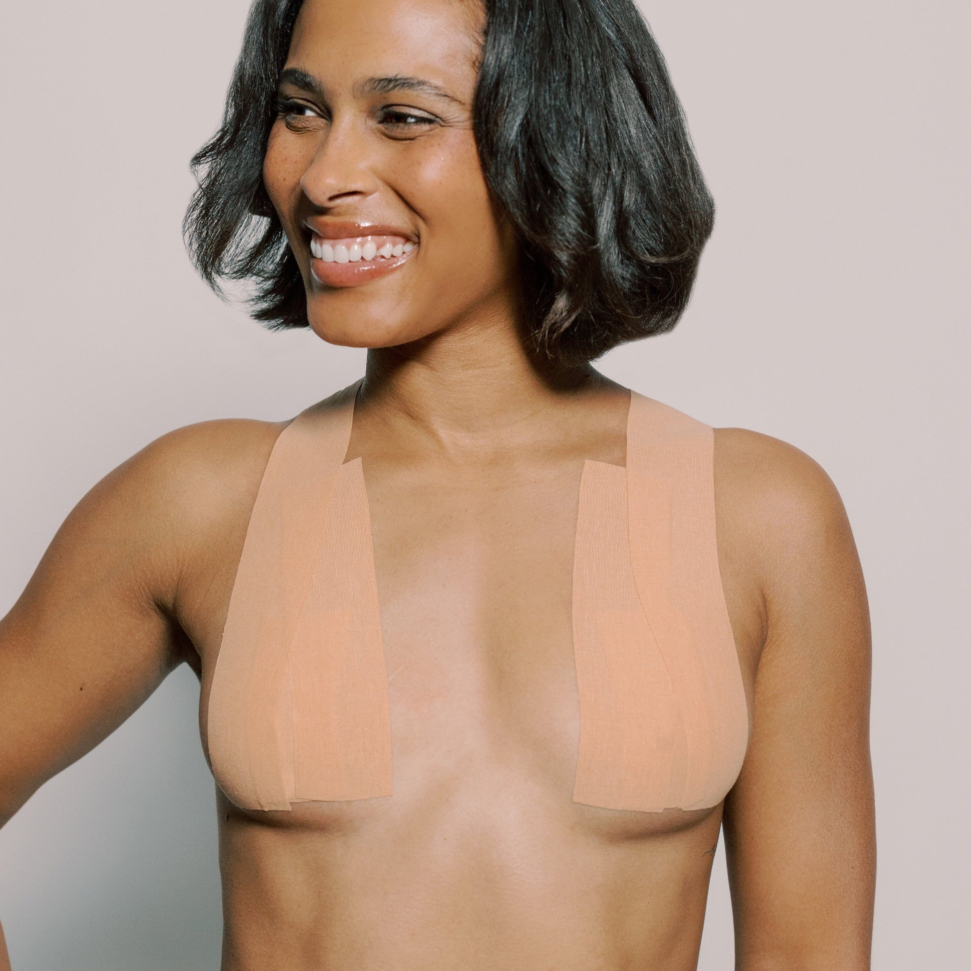 Model wears a few strips of tape from the Tape It Your Way breast tape roll. They are placed vertically and up on her shoulders showing how the tape can lift and shape. They are two separate strips, leaving plenty of space in the center of the chest for plunging necklines. Tape is nude in color.