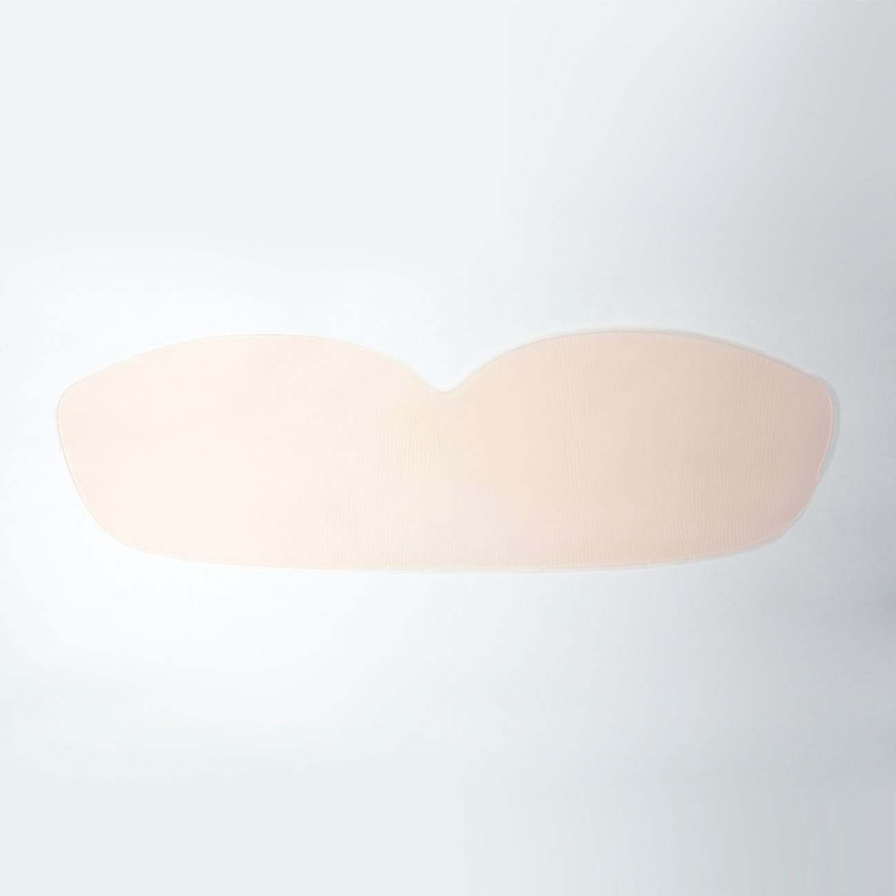 Silicone skin bandeau laid out flat on a surface to show its shape. It is long with a slightly curves bottom, tapered sides, and curved top that is shaped like a bra with the two curves being where each breast would be. Bra is transparent nude.