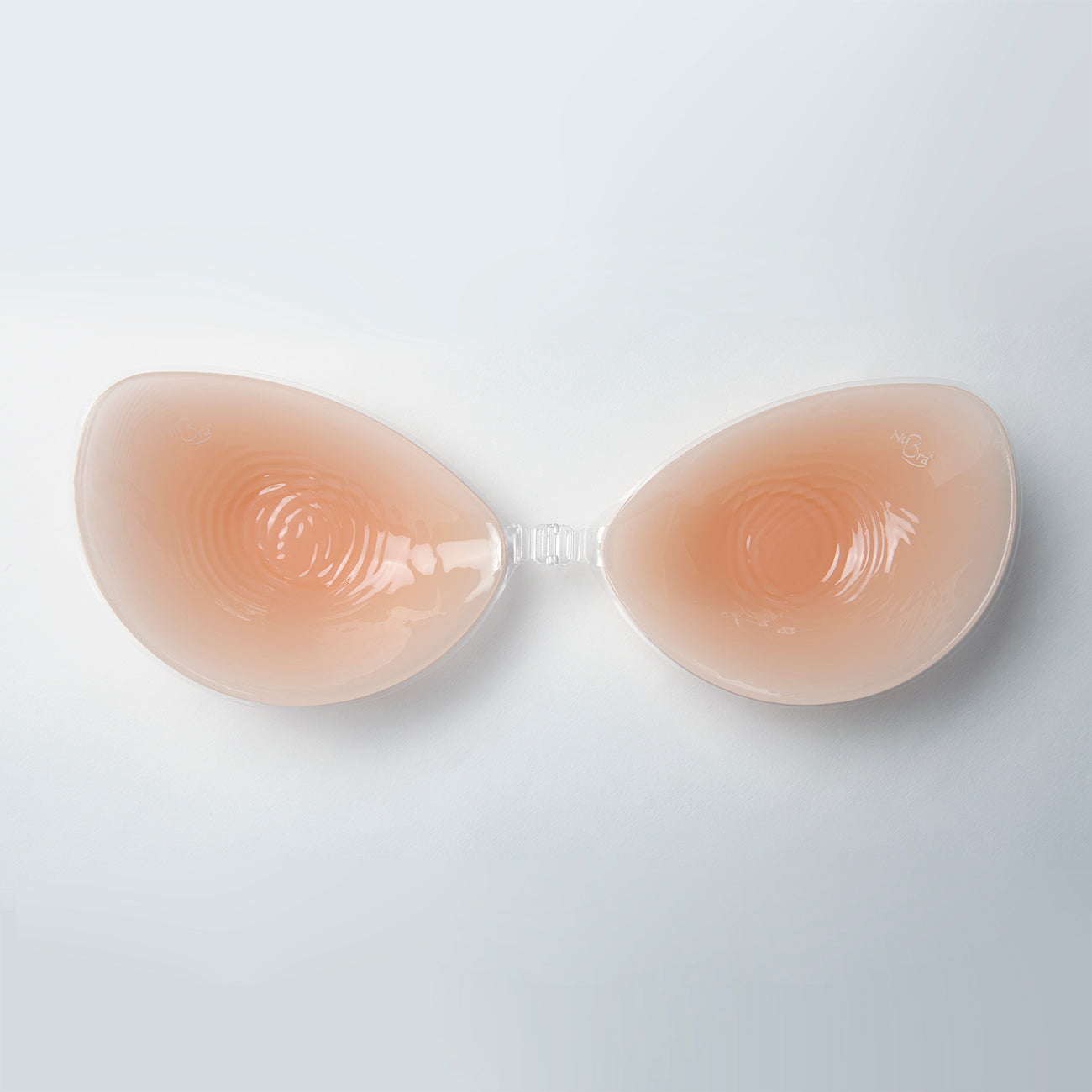 Close up of the nubra on its own. It is facing down, inside of cups facing the viewer. You can see how it will adhere to the body and where it clips in the middle. bra is nude in color.